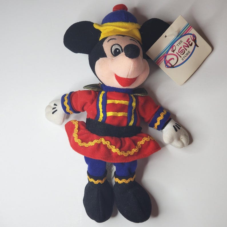 Puede incluir: Un mu&ntilde;eco de peluche de Mickey Mouse con un uniforme rojo y azul con adornos dorados, un sombrero a juego y guantes blancos. El mu&ntilde;eco tiene una cara sonriente y una etiqueta de color crema con el texto The Disney Store en azul y rojo.
