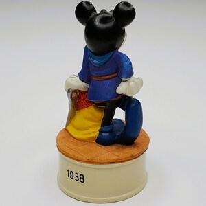 May include: A ceramic figurine of Mickey Mouse from the back, wearing a blue outfit and standing on a round base. The base is cream-colored with the year "1938" printed on it. The figurine has black ears and white gloves.