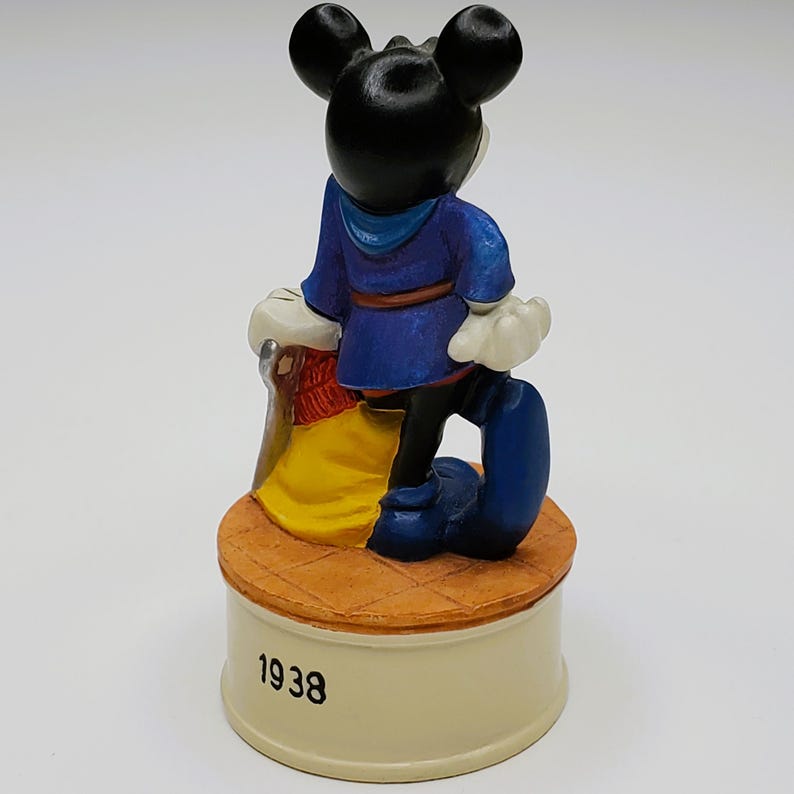 May include: A ceramic figurine of Mickey Mouse from the back, wearing a blue outfit and standing on a round base. The base is cream-colored with the year "1938" printed on it. The figurine has black ears and white gloves.