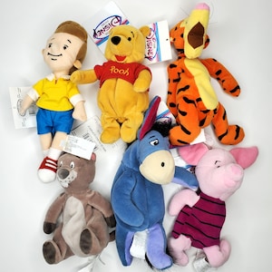 May include: A collection of six Disney Store plush toys including Christopher Robin in a yellow shirt and blue shorts, Winnie the Pooh in a red shirt, Tigger, a brown gopher, Eeyore, and Piglet wearing a striped shirt, all with attached tags.