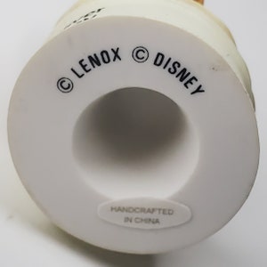 May include: A white ceramic figurine base with the text "LENOX" and "DISNEY" printed in black. The base has a circular shape with a hollow center. The words "HANDCRAFTED IN CHINA" are printed on a white sticker.