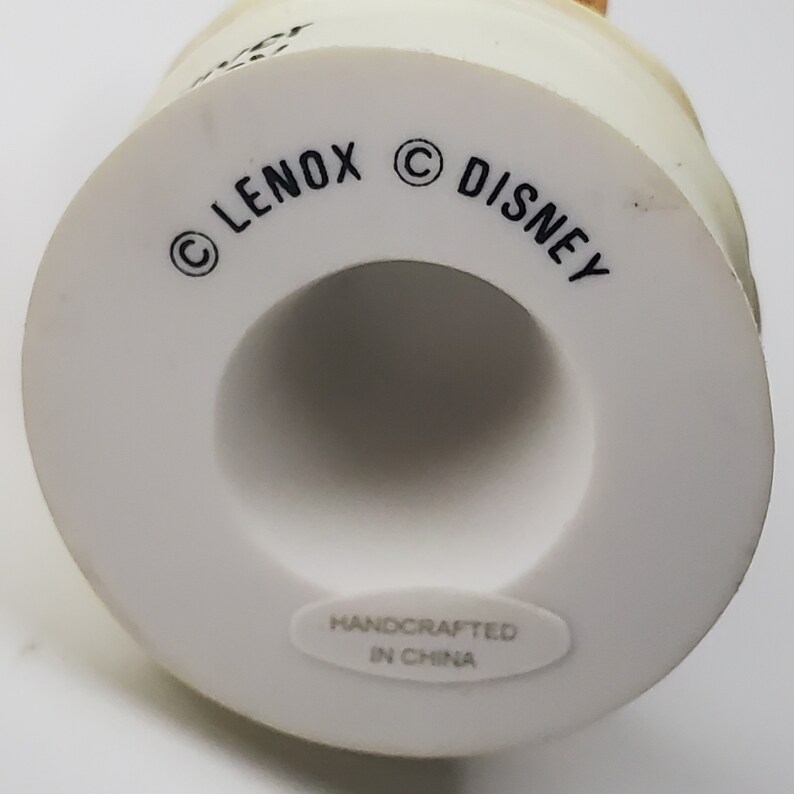 May include: A white ceramic figurine base with the text "LENOX" and "DISNEY" printed in black. The base has a circular shape with a hollow center. The words "HANDCRAFTED IN CHINA" are printed on a white sticker.