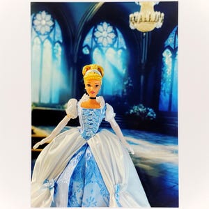 May include: A doll with blonde hair styled in an updo, wearing a light blue and white ball gown with a corset-style bodice, puffed sleeves, and white gloves. The doll features a black choker necklace and a blue headband, standing in a formal pose.
