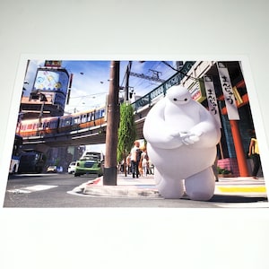 May include: A print featuring Baymax from Big Hero 6 standing on a city street. The image shows a train on an elevated track, buildings, and pedestrians. The Disney logo is in the bottom left corner.