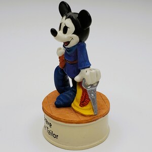 May include: A figurine of Mickey Mouse in a blue outfit, holding a saw. The figure stands on a round, tan base with the words "Brave Tailor" on the side. The base is set on a cream-colored pedestal.