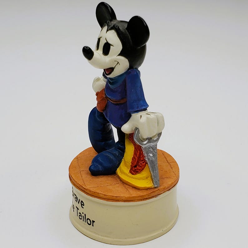 May include: A figurine of Mickey Mouse in a blue outfit, holding a saw. The figure stands on a round, tan base with the words "Brave Tailor" on the side. The base is set on a cream-colored pedestal.