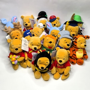 May include: A collection of yellow Winnie the Pooh plush toys in various costumes, including a bee, a graduate, a sailor, and a reindeer. Some wear hats, others hold flowers, and one is dressed as Tigger. Each plush features embroidered details.