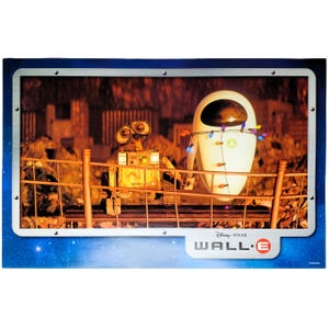 May include: A rectangular print featuring the characters WALL-E and EVE from the Disney Pixar film WALL-E. The robots stand behind a metal fence, with EVE draped in colorful string lights. The frame has a metallic border and the movie logo.