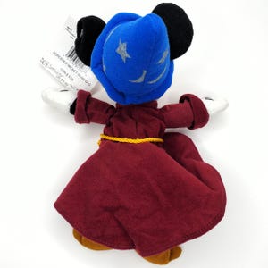 May include: A Sorcerer Mickey Mouse bean bag plush toy. The plush has a blue sorcerer hat with silver stars, a burgundy robe, and yellow rope belt. The toy has black ears, white gloves, and brown shoes. The tag reads "SORCERER MICKEY BEAN BAG".