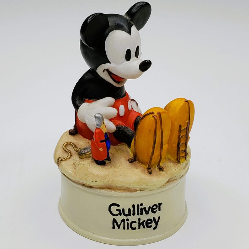 May include: A figurine of Mickey Mouse in a seated position, wearing red shorts, white gloves, and yellow shoes. The figurine is on a base with the words "Gulliver Mickey" and includes a small figure of a person in a red coat.