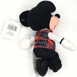 May include: A Mickey Mouse soft toy, dressed in a red, black, and blue tartan waistcoat, black trousers, and black shoes. The toy has black ears, a black head, and white gloves. A tag reads "Mickey Tour Guide Mini Bean Bag."