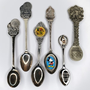 May include: A collection of vintage souvenir spoons featuring various designs. The spoons showcase themes like Epcot Center, Walt Disney World, and Animal Kingdom. The spoons are silver-toned and have detailed embellishments, including character figures and architectural elements.