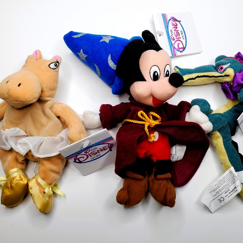 K&ouml;nnte beinhalten: Eine Sammlung von Disney-Pl&uuml;schtieren, darunter Mickey Mouse als Zauberer, ein Ballerina-Nilpferd und ein gr&uuml;nes Alligator. Jedes Spielzeug hat ein Etikett von The Disney Store. Das Mickey Mouse-Pl&uuml;schtier tr&auml;gt einen roten Umhang und einen blauen Zaubererhut.
