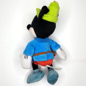 May include: A plush toy of a cartoon character, featuring a black head and ears, a blue shirt, red pants, and light blue shoes. The character wears a green hat and has white gloves. The toy is on a white background.