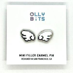 Puede incluir: Un par de pines de esmalte blanco pixelado con forma de ala y bordes plateados. Los pines se muestran en una tarjeta blanca con el texto "Olly Bits" en morado y azul, y "Mini Filler Enamel Pin" y "Designed in San Francisco, CA" en negro.