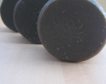 May include: Three round, dark gray soap bars with a textured surface. The bar in the foreground has a small, stylized flower design.