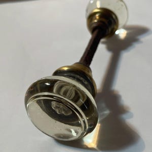 May include: A vintage glass door knob with a clear, rounded design. The knob has a brass base and a dark brown stem. The glass is clear with a slight tint, and the knob is likely from an antique door.