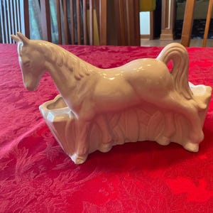May include: A cream-colored ceramic horse planter. The horse is in a trotting pose, with its head lowered and tail curved. The planter has a rectangular base with a trough for plants. The horse is detailed with a mane and tail.