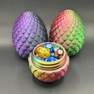 Op de afbeelding: Een paars-groene drakenei-vormige dobbelstenenhouder met een deksel dat opengaat om een ​​set kleurrijke dobbelstenen te onthullen. De dobbelstenen zijn een mix van blauw, geel en rood.