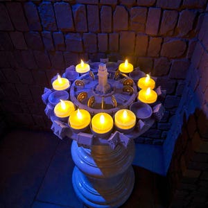 Op de afbeelding: Een decoratieve kandelaar met een centraal torenontwerp, omringd door een cirkelvormige opstelling van brandende, gele LED-kaarsen. De houder wordt ondersteund door een witte, sierlijke sokkel. De achtergrond toont een getextureerde, donkere stenen muur.