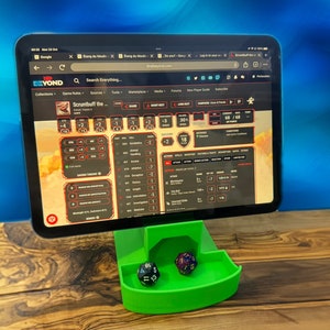 Op de afbeelding: Een groene plastic dobbelsteenbak met twee dobbelstenen, een zwarte en een groene, op een houten tafel. Een tablet met de website "D&D Beyond" staat op een standaard achter de dobbelsteenbak. De website toont een personageblad met de naam "Scruntbuff the..." en verschillende statistieken.