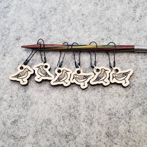 May include: Eight wooden stitch markers with a bird design, attached to a wooden knitting needle. The markers are brown and have black bird silhouettes.
