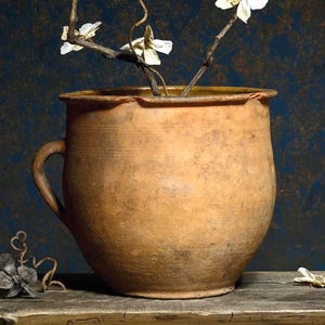 May include: A still life image showing a terracotta pot with a handle, containing white flowers on thin stems. The pot rests on a weathered wooden surface, set against a dark, textured background.
