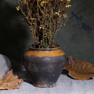 May include: A rustic still life arrangement. A dark, aged ceramic pot with a yellow band holds dried yellow flowers. A large, dried brown leaf and a grey cloth are in the foreground, set against a dark green backdrop.