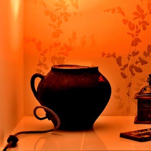 Pode incluir: Um pote de cerâmica castanho-escuro com pega, um pequeno moinho de café de madeira, um frasco de vidro com fecho metálico, uma tablete de chocolate retangular e um saco de papel bege sob luz laranja quente numa prateleira quadrada.