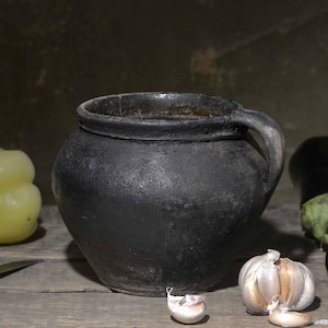 Op de afbeelding: Een donkere, getextureerde keramische pot met één handvat staat centraal, omringd door een lichtgele paprika, twee donkerpaarse aubergines, een bol knoflook met losse teentjes en een metalen mes met een houten handvat op een houten ondergrond.