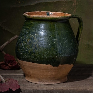 May include: A rustic, green and tan ceramic pitcher with a handle, set on a wooden surface. The pitcher has a textured surface and a wide base. Dried branches and red leaves are in the foreground.