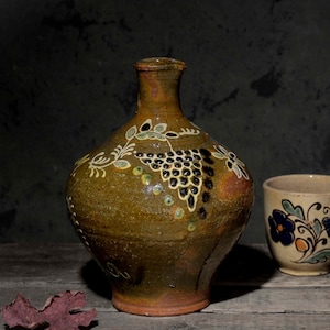 May include: A brown ceramic jug with a narrow neck and a hand-painted grape cluster design in white, green, and black, paired with a cream-colored ceramic mug featuring a hand-painted floral pattern in blue, green, and orange tones.