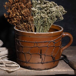 Op de afbeelding: Een rustig stilleven met een bruine keramische mok gevuld met gedroogde bloemen, een gevouwen linnen doek en een walnoot op een houten oppervlak. De mok heeft een draadkooi-ontwerp. Een klein mes ligt in de buurt.