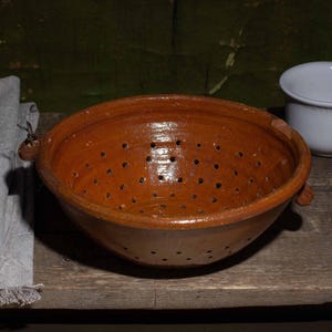May include: A rustic kitchen scene featuring a brown ceramic colander with drainage holes, a wooden spoon, a grey linen napkin, and a white ceramic cup. The items are arranged on a weathered wooden surface.