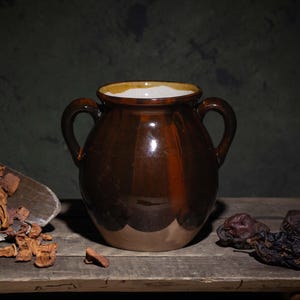 May include: A still life composition featuring a brown ceramic jar with two handles, filled with a white substance. A wooden scoop holds dried fruit, and a few pieces are scattered on the rustic wooden surface. Dark background.