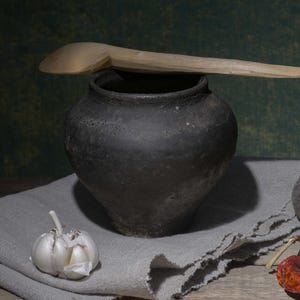 Puede incluir: Naturaleza muerta rústica con una olla de barro oscura y una cuchara de madera. La escena incluye ajo, remolacha y un pimiento rojo seco sobre una superficie de madera, con un paño de lino gris.