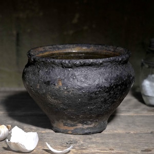 May include: A dark, textured, cast iron pot with a wide rim and rounded body, accompanied by a whole bulb of garlic, several loose garlic cloves, and a clear glass jar containing a white granular substance, all arranged on a wooden surface.