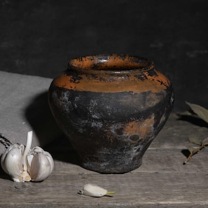 Puede incluir: Naturaleza muerta rústica con una olla de cerámica oscura y desgastada con una banda naranja, un paño de lino gris y un bulbo de ajo. La composición está sobre una superficie de madera, con un fondo oscuro.