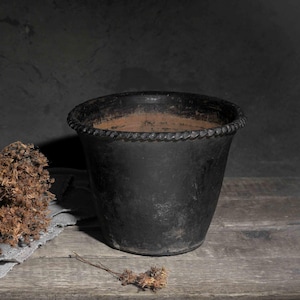 May include: A black, textured plant pot with a rope-style rim on a wooden surface. A bundle of dried brown flowers rests on a grey linen cloth. The background is a dark, textured wall.