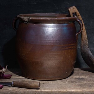 May include: A rustic still life featuring a large brown ceramic pot with handles, a wooden spoon, a red onion, garlic, and a knife. The items are arranged on a wooden surface against a dark background, evoking a sense of traditional cooking.