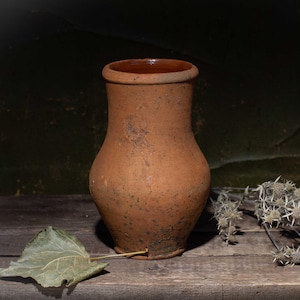 Puede incluir: Una jarra rústica de barro marrón con base ancha y cuello estrecho sobre una superficie de madera envejecida. Una hoja seca y ramitas de flores secas dispuestas cerca. El fondo es una pared oscura y texturizada.