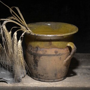 Op de afbeelding: Een rustieke keramische pot met een mosterdgele geglazuurde rand en een verweerde bruine behuizing, voorzien van een handvat aan de zijkant. Verschillende stengels gedroogde gouden tarwe hangen over de rand, naast een stuk donkergrijze leisteen.