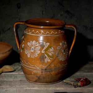 May include: A brown terracotta pitcher with two handles and hand-painted white and green floral motifs, accompanied by a matching terracotta colander, a wooden spoon, and a single dried red chili pepper arranged on a wooden surface.