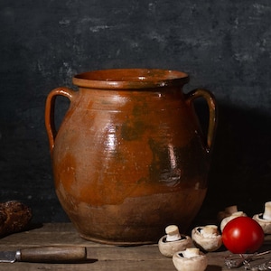 May include: A still life composition featuring a large, brown earthenware pot with handles, a loaf of bread, a knife, mushrooms, and a red tomato. The items are arranged on a wooden surface against a dark background, evoking a rustic, culinary theme.