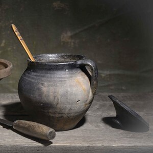 Op de afbeelding: Een donkergrijze keramische pot met een handvat, een houten lepel erin, een bruine keramische kom, een hele gele ui, een metalen mes met een houten handvat en een klein zwart dekselstuk op een houten ondergrond gerangschikt.