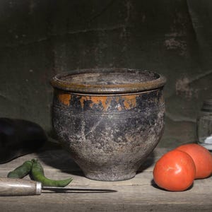 Puede incluir: Una vasija de cerámica desgastada de color gris oscuro y marrón se sitúa en el centro, rodeada por una berenjena morada, dos chiles verdes, un cuchillo de metal con mango de madera, dos tomates rojos, un frasco de sal y un pimiento amarillo.