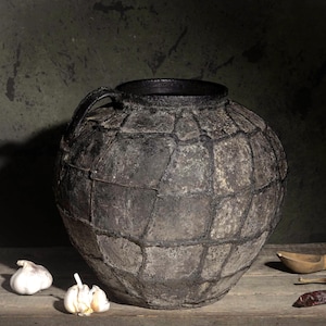 May include: A large, round, dark gray ceramic vessel with a textured, grid-like surface pattern and a single handle. Two white garlic bulbs sit to the left, while a wooden spoon and two dried red chili peppers are positioned to the right.