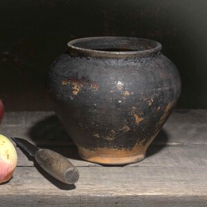 Op de afbeelding: Een donkere, verweerde keramische pot met een wijde opening en een ronde vorm staat naast drie verse appels en een klein schilmesje. De appels vertonen tinten rood, geel en groen, waarbij één appel een klein, gedroogd blaadje aan de steel heeft.