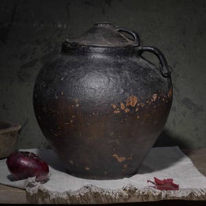 Op de afbeelding: Een grote, ronde, donkerbruine en zwarte keramische kruik met een deksel en een handvat aan de zijkant. Het oppervlak heeft een getextureerd, verweerd uiterlijk met zichtbare slijtageplekken. Een kleine houten kom en een rode ui staan ernaast.