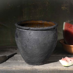 Może przedstawiać: Ciemny, teksturowany, matowy czarny garnek ceramiczny z szerokim brzegiem, umieszczony obok małej pomarańczowej miski zawierającej plaster czerwonego arbuza, oddzielny kawałek arbuza oraz metalowe narzędzie z ciemną drewnianą rączką.